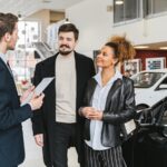 Salesperson speaking with two customers beside cars in a dealership